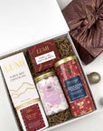 A curated gift box set containing a box of Lumi White Hot Chocolate, a jar of Whittard marshmallows, and a tin of Whittard Orange Grands & Dark Chocolates, a candle, box of lumi caramels, arranged in a white box with brown crinkle paper filler, alongside a wrapped gift and holiday ornaments.