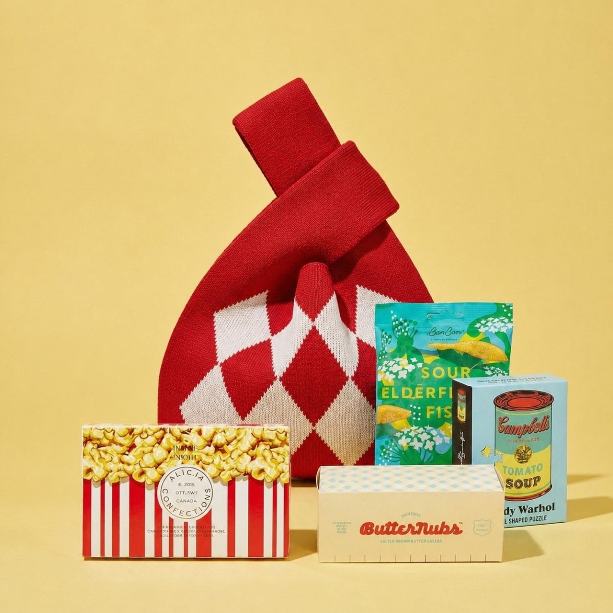 A curated gift set on a solid pale yellow background, featuring a red and white argyle knit bag, a popcorn box, a ButterNub's box, an Andy Warhol Campbell's Soup box, and Bon Bon Sour Elderfish packet.