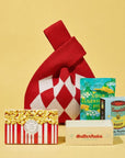 A curated gift set on a solid pale yellow background, featuring a red and white argyle knit bag, a popcorn box, a ButterNub's box, an Andy Warhol Campbell's Soup box, and Bon Bon Sour Elderfish packet.