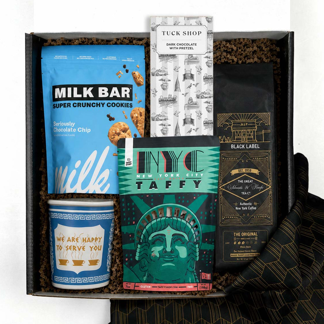 A curated gift box set containing a blue bag of Milk Bar cookies, a bag of coffee with a black and gold label, a green NYC Taffy box, a blue anthora we are happy to serve you ceramic cup, a Tuck Shop chocolate bar, and a black patterned fabric napkin, all arranged in a box with brown crinkle paper. 