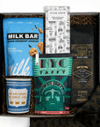 A curated gift box set containing a blue bag of Milk Bar cookies, a bag of coffee with a black and gold label, a green NYC Taffy box, a blue anthora we are happy to serve you ceramic cup, a Tuck Shop chocolate bar, and a black patterned fabric napkin, all arranged in a box with brown crinkle paper. 