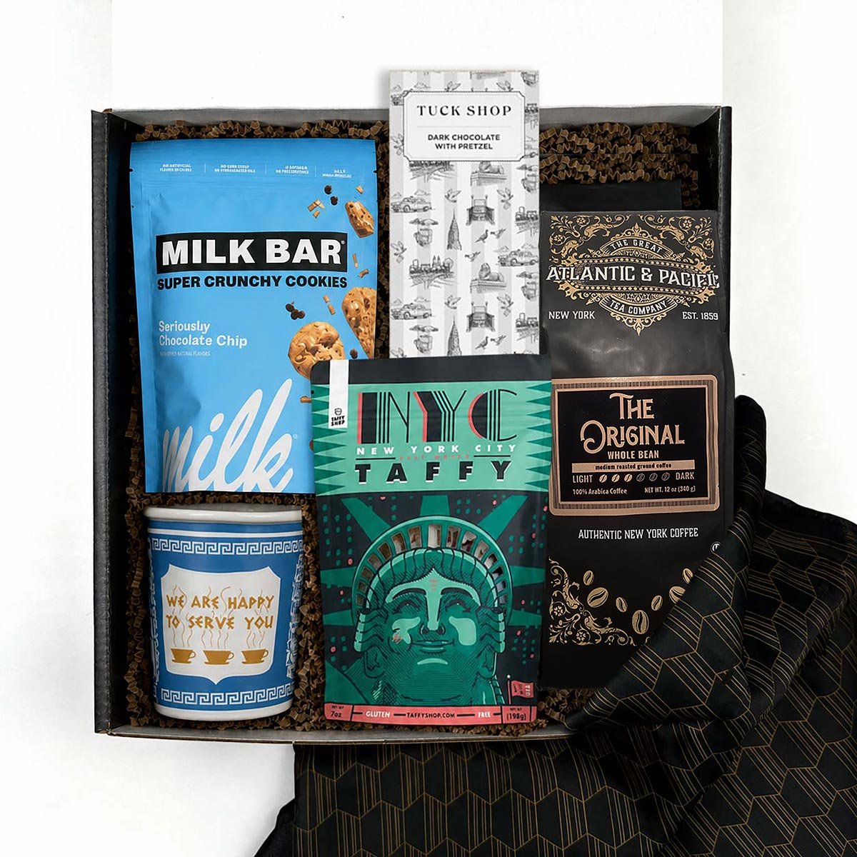 A curated gift box set containing a blue bag of Milk Bar cookies, a bag of coffee with a black and gold label, a green NYC Taffy box, a blue anthora we are happy to serve you ceramic cup, a Tuck Shop chocolate bar, and a black patterned fabric napkin, all arranged in a box with brown crinkle paper. 