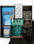 A curated gift box set containing a blue bag of Milk Bar cookies, a bag of coffee with a black and gold label, a green NYC Taffy box, a blue anthora we are happy to serve you ceramic cup, a Tuck Shop chocolate bar, and a black patterned fabric napkin, all arranged in a box with brown crinkle paper. 