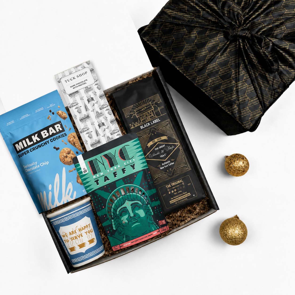 A curated gift box assortment on a white background, featuring a milk bar cookie tin, a chocolate bar, a bag of coffee, a taffy package, and a black patterned fabric wrapped gift box.