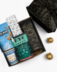 A curated gift box assortment on a white background, featuring a milk bar cookie tin, a chocolate bar, a bag of coffee, a taffy package, and a black patterned fabric wrapped gift box.