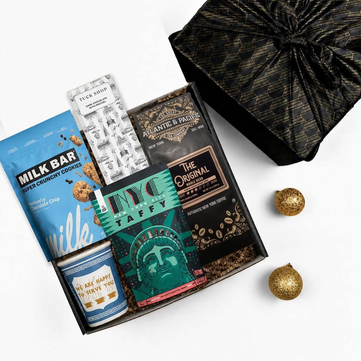 A curated gift box assortment on a white background, featuring a milk bar cookie tin, a chocolate bar, a bag of coffee, a taffy package, and a black patterned fabric wrapped gift box.