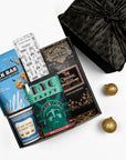 A curated gift box assortment on a white background, featuring a milk bar cookie tin, a chocolate bar, a bag of coffee, a taffy package, and a black patterned fabric wrapped gift box.