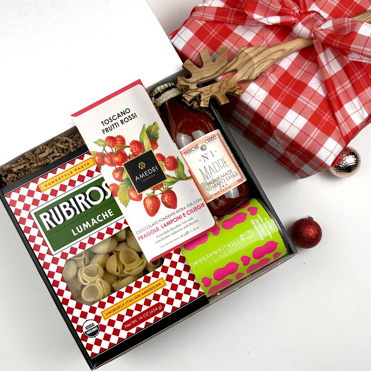 kadoo pasta holiday gift box with red tartan furoshiki wrap. Inside gifts rubirosa lumache pasta, amedei toscano frutti chocolate bar, madre tomato sauce, can of tomato and more