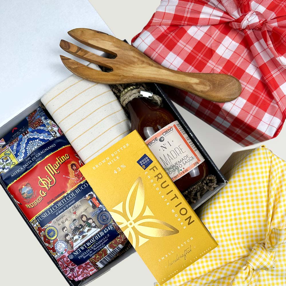 kadoo pasta night curated gift box in red fabric wrap with dry pasta, chocolate bar, homemade tomato sauce, yellow stripes kitchen towel, olive wooden fork utensil, and more. It has red dried flower bouquet.