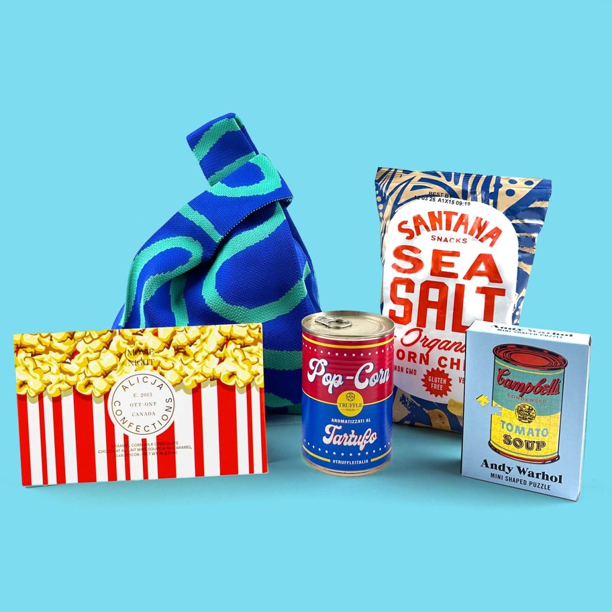 Collection of snack items including a blue bag, popcorn box, truffle popcorn, sea salt chips, and Andy Warhol tomato soup can puzzle on a blue background.