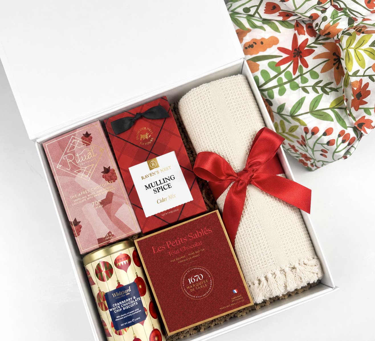 A curated gift box set containing a cream knit blanket tied with a red ribbon, a tin of Whittard cranberry white chocolate chip biscuits, a box of Les Petits Sables cookies, and Ritual Apres chocolate bar, all arranged in a white box next to a floral fabric gift wrap.