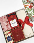A curated gift box set containing a cream knit blanket tied with a red ribbon, a tin of Whittard cranberry white chocolate chip biscuits, a box of Les Petits Sables cookies, and Ritual Apres chocolate bar, all arranged in a white box next to a floral fabric gift wrap.