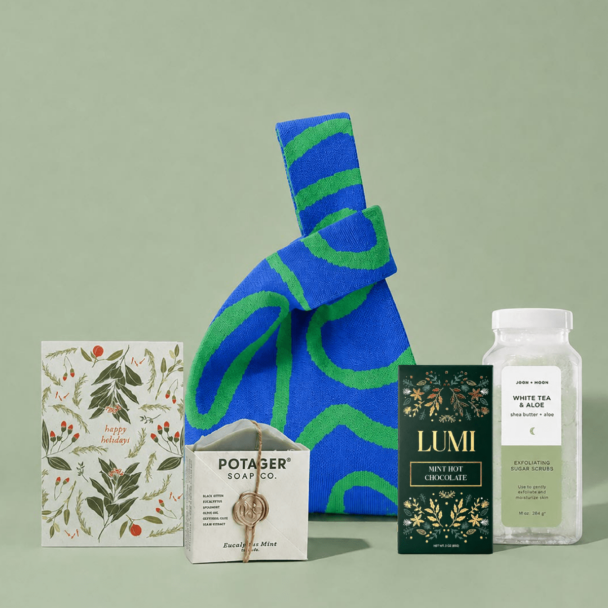 A curated gift set arranged on a pale green background, featuring a blue and green patterned fabric tote bag, a botanical card, a box of Potager Soap Co. soap, a Lumi Hot chocolate box, and a bottle of tea.