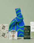 A curated gift set arranged on a pale green background, featuring a blue and green patterned fabric tote bag, a botanical card, a box of Potager Soap Co. soap, a Lumi Hot chocolate box, and a bottle of tea.