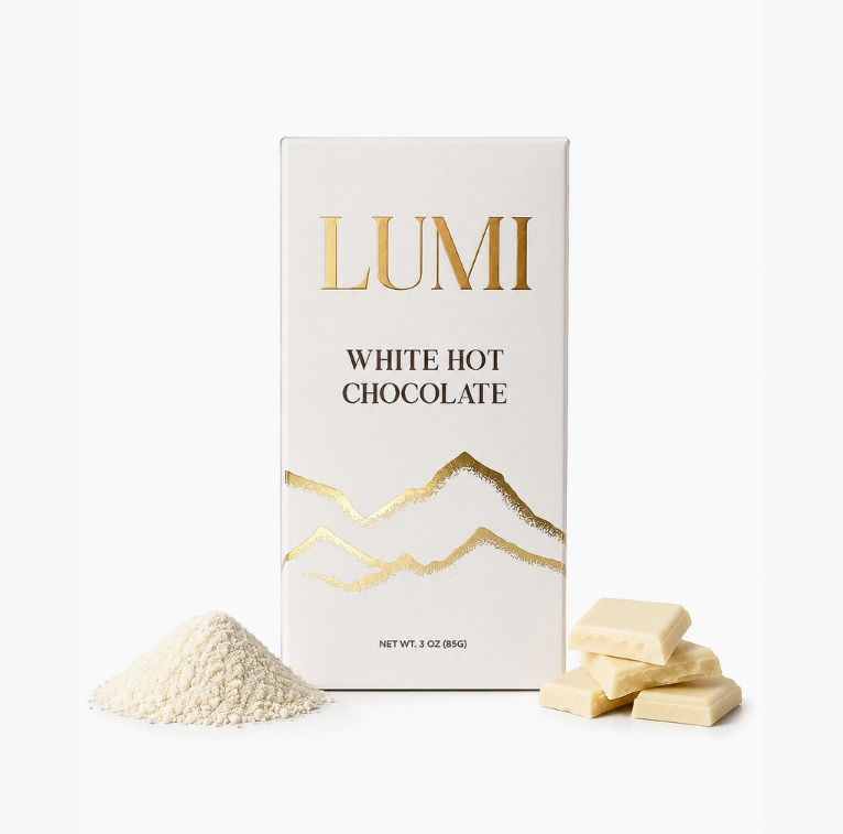 A box of Lumi White Hot Chocolate shown on a clean white background, with a small pile of white hot chocolate powder and several white chocolate pieces placed in front of the package.