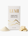 A box of Lumi White Hot Chocolate shown on a clean white background, with a small pile of white hot chocolate powder and several white chocolate pieces placed in front of the package.
