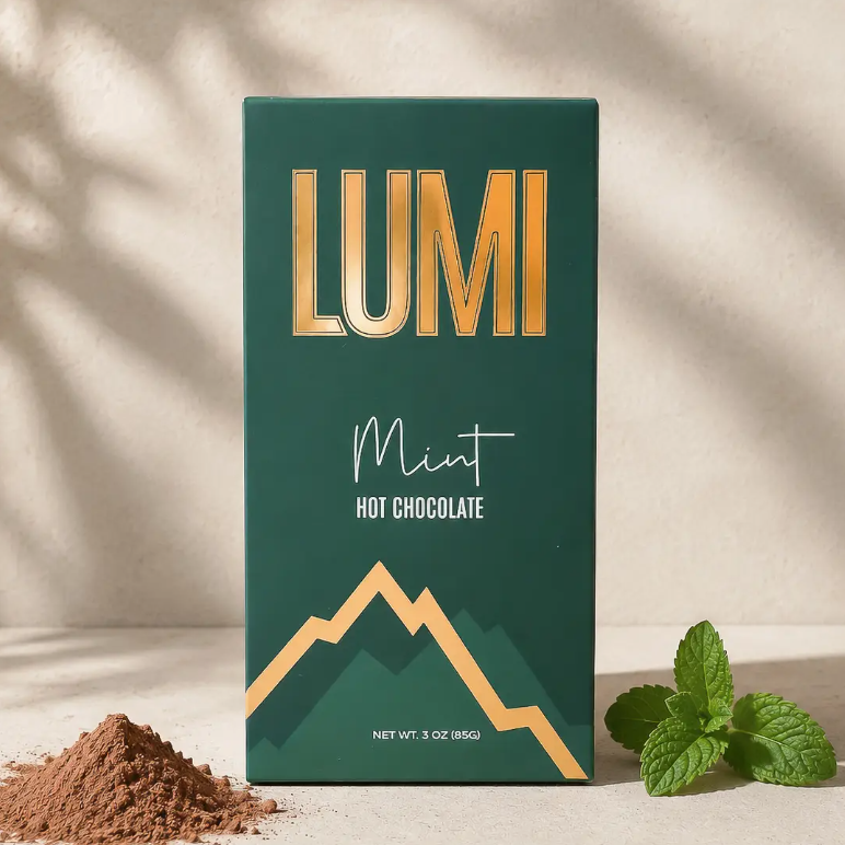 A 3 oz box of Lumi Mint Hot Chocolate, featuring a dark green box with gold lettering and a mountain graphic, styled with cocoa powder and fresh mint leaves on a sunlit beige surface.