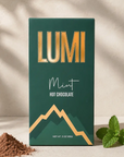 A 3 oz box of Lumi Mint Hot Chocolate, featuring a dark green box with gold lettering and a mountain graphic, styled with cocoa powder and fresh mint leaves on a sunlit beige surface.