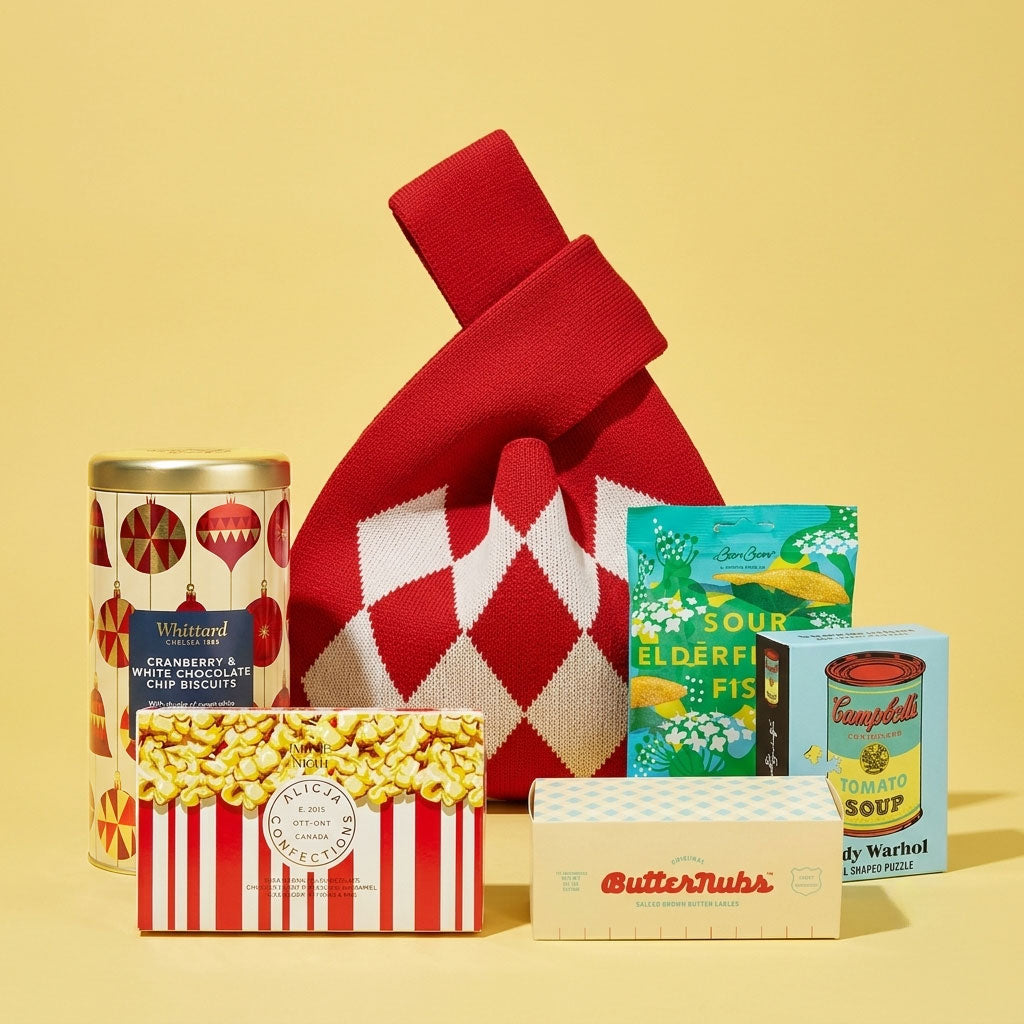 A curated gift bag arrangement on a pale yellow background, featuring a red-and-white argyle knit bag, a can of Whittard cranberry and white chocolate chip cookies, a milk chocolate bar by alicja confections, a box of ButterNubs Brown Butter Sablé Cookies , a packet of BonBon sour elderfish, and a box of Andy Warhol Campbell's tomato soup puzzle.
