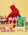 A curated gift bag arrangement on a pale yellow background, featuring a red-and-white argyle knit bag, a can of Whittard cranberry and white chocolate chip cookies, a milk chocolate bar by alicja confections, a box of ButterNubs Brown Butter Sablé Cookies , a packet of BonBon sour elderfish, and a box of Andy Warhol Campbell's tomato soup puzzle.