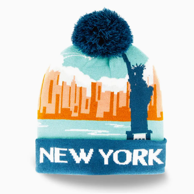 A knit New York beanie hat featuring a navy blue pom-pom, a light blue and orange city skyline graphic with the Statue of Liberty, and a navy blue cuff with 'NEW YORK' in white text.