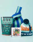 A New York City themed gift set on a light blue background, featuring a bag of NYC Statue of Liberty taffy, a small box of French Chocolate with NYC theme art print, and a blue knit beanie of a New York City skyline with a pom-pom.
