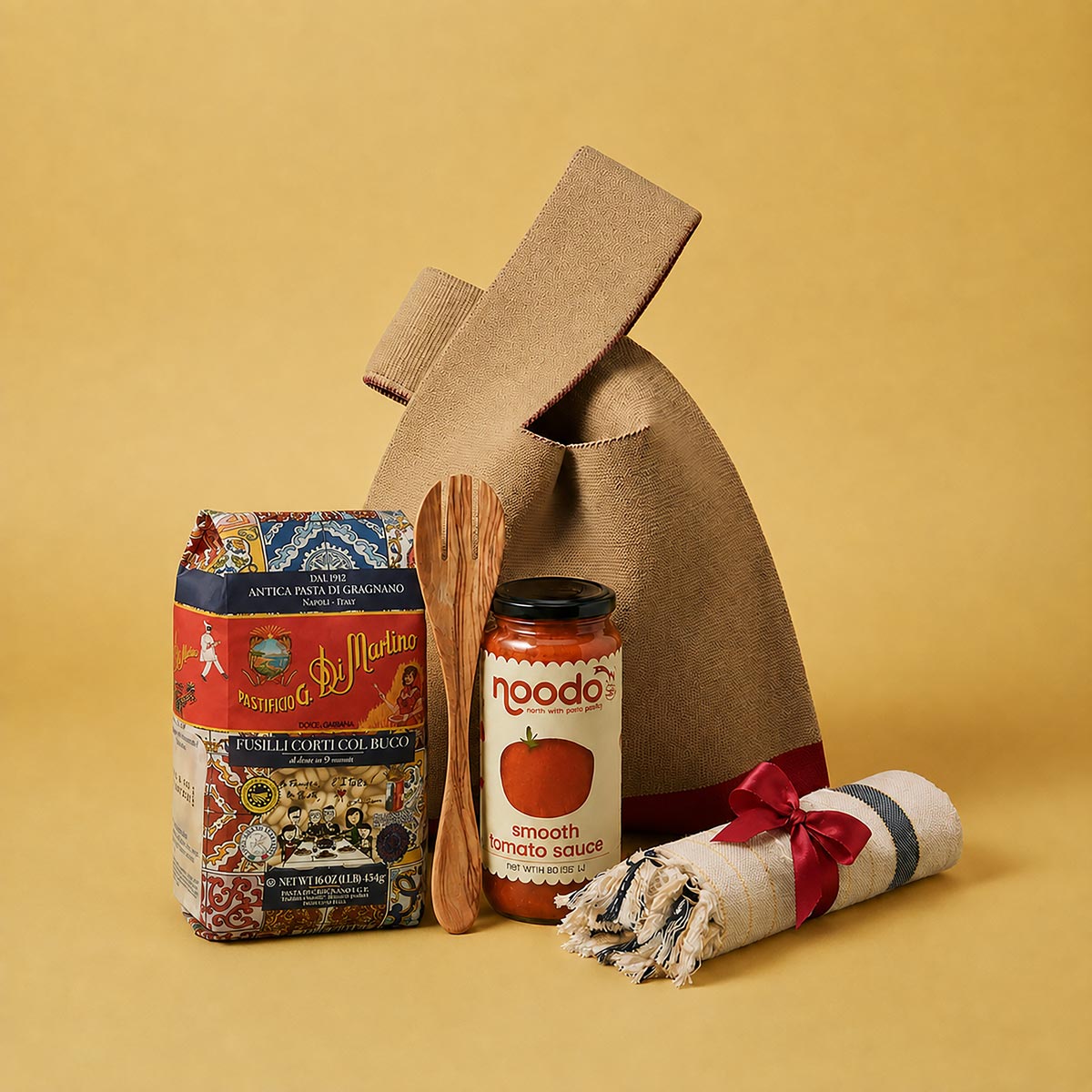 A gift bag set featuring a brown canvas bag, a jar of noodo smooth tomato sauce, a bag of Fusili Corti Pasta, a wooden spoon, and a rolled white towel with a red ribbon, arranged on a solid yellow background.