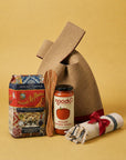 A gift bag set featuring a brown canvas bag, a jar of noodo smooth tomato sauce, a bag of Fusili Corti Pasta, a wooden spoon, and a rolled white towel with a red ribbon, arranged on a solid yellow background.