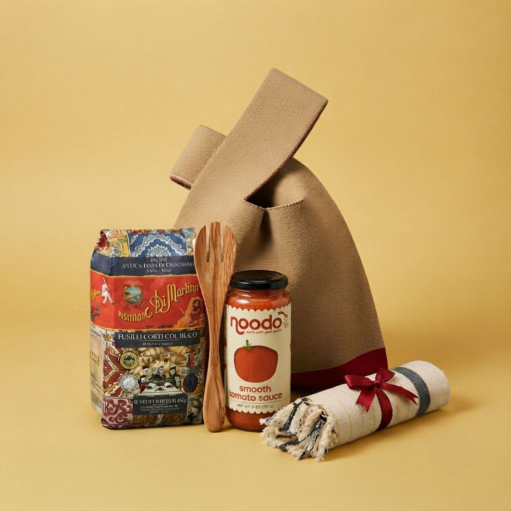 A gift bag set featuring a brown canvas bag, a jar of noodo smooth tomato sauce, a bag of Fusili Corti Pasta, a wooden spoon, and a rolled white towel with a red ribbon, arranged on a solid yellow background.