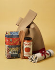 A gift bag set featuring a brown canvas bag, a jar of noodo smooth tomato sauce, a bag of Fusili Corti Pasta, a wooden spoon, and a rolled white towel with a red ribbon, arranged on a solid yellow background.
