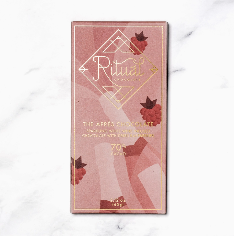 A single bar of Ritual The Apres chocolates in its dusty pink and rose gold packaging with gold foil details and illustrations of rasberries and wine bottles, set against a white marble background.