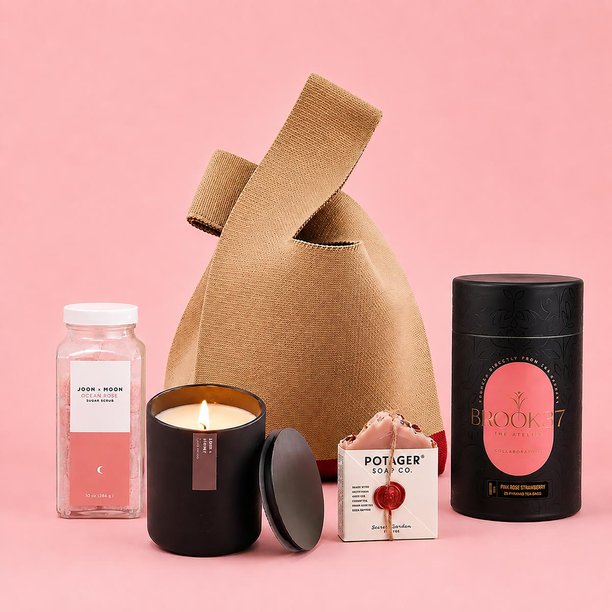 A curated gift bag set arranged on a pink studio background, featuring a beige knit bag, a black Rose + Peony fragrant candle jar with a lit candle and lid beside it, a clear bottle of pink Exfoliating Sugar Cubes by Joon x Moon, a small pink hand made potager soap, and a black cylindrical container of Refreshing Pink Rose Strawberry Tea by Brook37 Atelier.