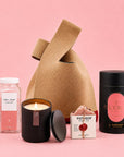 A curated gift bag set arranged on a pink studio background, featuring a beige knit bag, a black Rose + Peony fragrant candle jar with a lit candle and lid beside it, a clear bottle of pink Exfoliating Sugar Cubes by Joon x Moon, a small pink hand made potager soap, and a black cylindrical container of Refreshing Pink Rose Strawberry Tea by Brook37 Atelier.