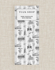 A bar of Tuck Shop Dark Chocolate bar in a white and gray striped packaging featuring black line-art illustrations of London landmarks on a textured beige background.