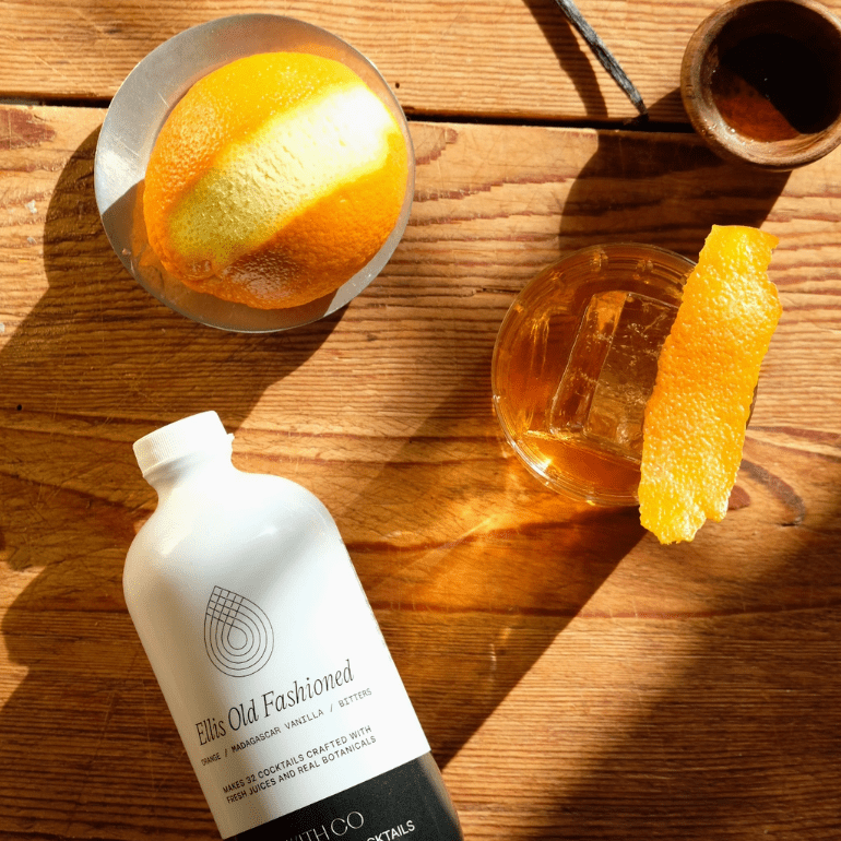 A bottle of WithCo Ellis Old Fashion Mix  lying on a rustic wooden table, styled with an orange slice in a glass, vanilla beans, and cinnamon sticks under warm, direct sunlight.