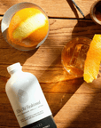 A bottle of WithCo Ellis Old Fashion Mix  lying on a rustic wooden table, styled with an orange slice in a glass, vanilla beans, and cinnamon sticks under warm, direct sunlight.